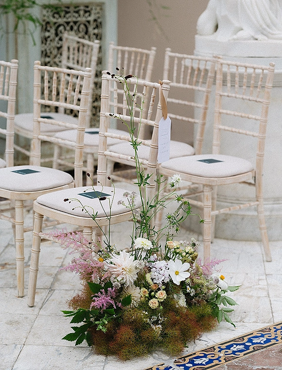 Ceremony aisle decor with wedding aisle flower arrangement and greenery on a stone floor beside chiavari chairs with chair tags indoors