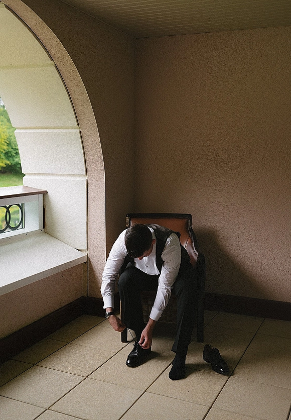Groom getting ready, tying shoe laces on black dress shoes while seated on a leather chair by an arched window and balcony railing