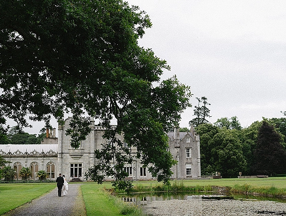 Couple walking away along a gravel path, wedding dress train trailing beside groom’s suit, with a stone manor house and pond behind