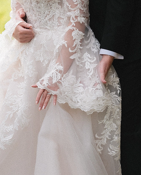 Wedding dress close-up with lace wedding dress sleeves and sheer floral appliqué as bride’s ringed hand rests on groom’s cuff outdoors