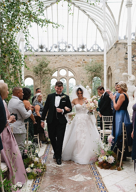 Wedding recessional with bride and groom walking aisle, her lace dress and veil flowing as guests cheer under a glass conservatory ceiling