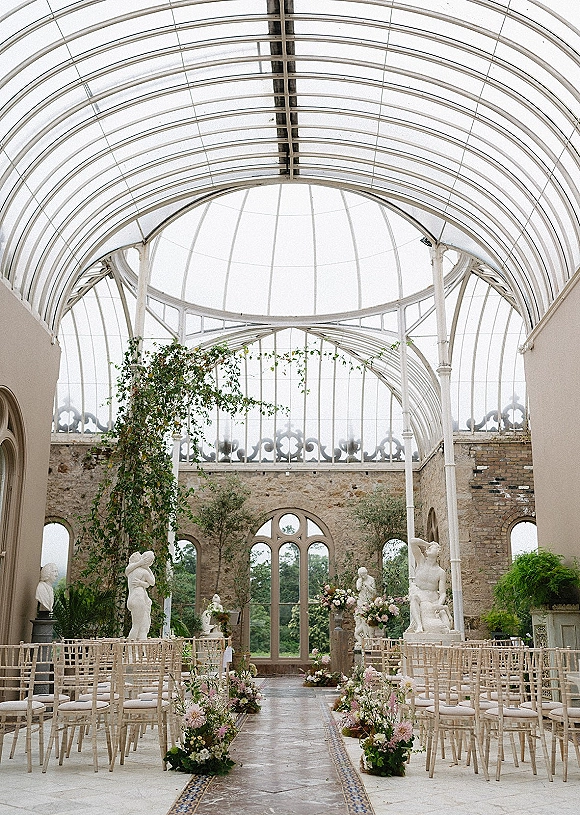 Ceremony setup with aisle florals lining a tiled runner between chiavari chairs under a glass conservatory ceiling and arched windows