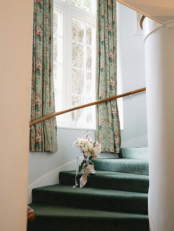 Bridal shoes with a wedding shoes on stairs display, paired with a pastel bouquet and ribbon on a green carpeted staircase by a window