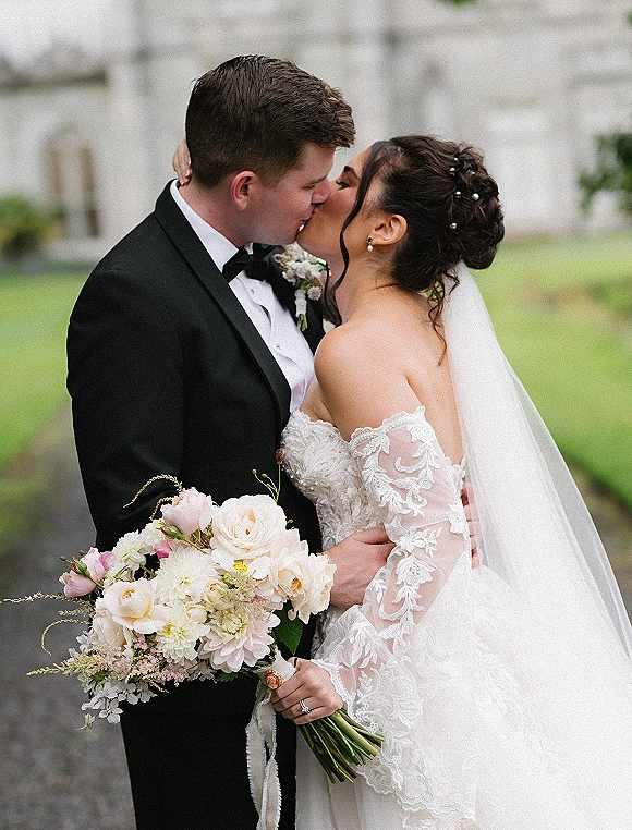 Wedding kiss portrait of bride and groom kiss, she holds a bouquet and veil on an outdoor lawn by trees and a stone building walkway