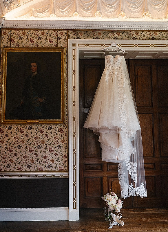 Wedding dress with lace wedding dress detailing hanging on a wood-paneled door, paired with veil, bouquet, ribbon, and heels in a vintage room