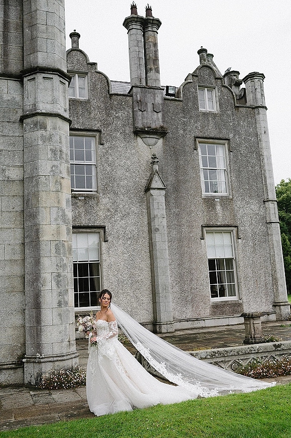 Bridal portrait of bride holding bouquet in an off-the-shoulder lace gown with cathedral veil trailing on lawn by stone manor terrace