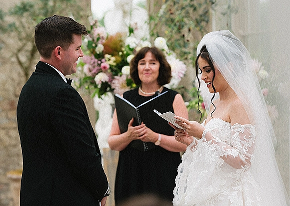 Wedding vows as bride reading vows from a booklet while groom listens, her cathedral veil flowing in an outdoor garden ceremony backdrop