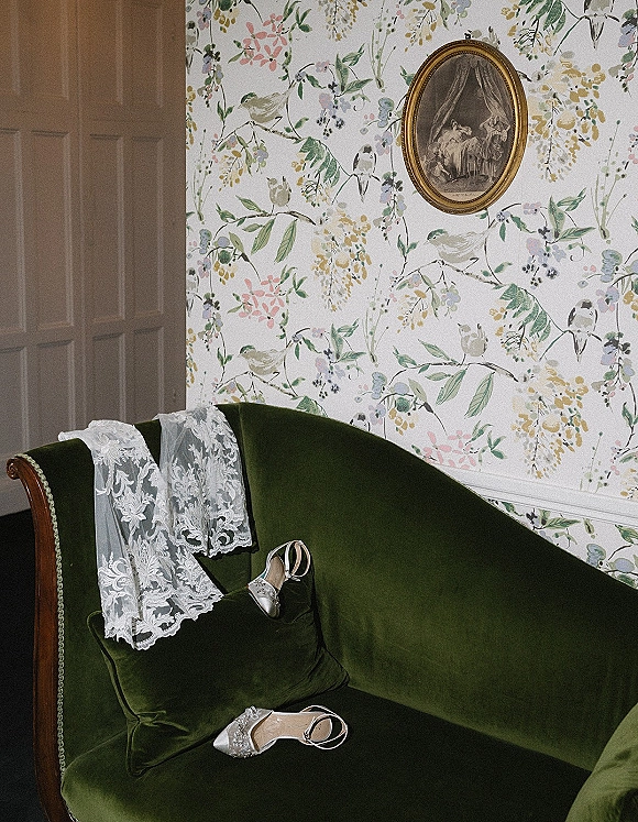 Bridal accessories with wedding shoes detail—satin ankle-strap heels and lace veil draped on a green velvet sofa against floral wallpaper