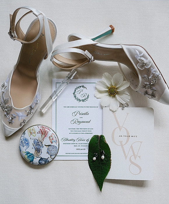 Invitation suite wedding invitation flat lay with white bridal shoes, vow card, perfume bottle, pearl earrings on leaf, on light fabric surface