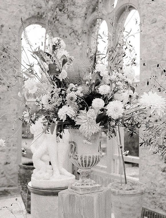Wedding floral arrangement in an urn wedding centerpiece with wildflowers and greenery on a pedestal beside a stone statue in a courtyard