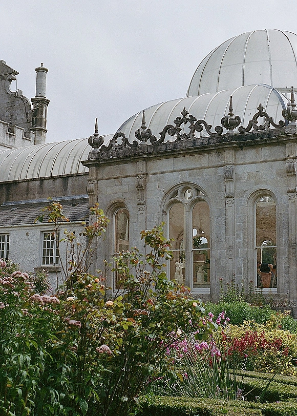 Wedding venue exterior with a stone conservatory facade, arched windows and a glass dome roof, framed by manicured hedges and flowers under overcast sky