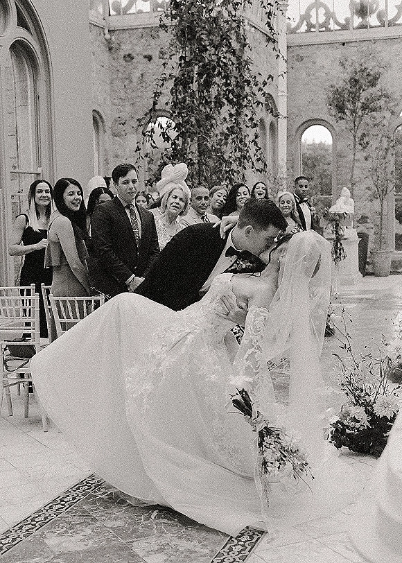 Wedding kiss moment as groom dips the bride in a lace long-sleeve gown, veil trailing, in a stone courtyard with guests and arched windows