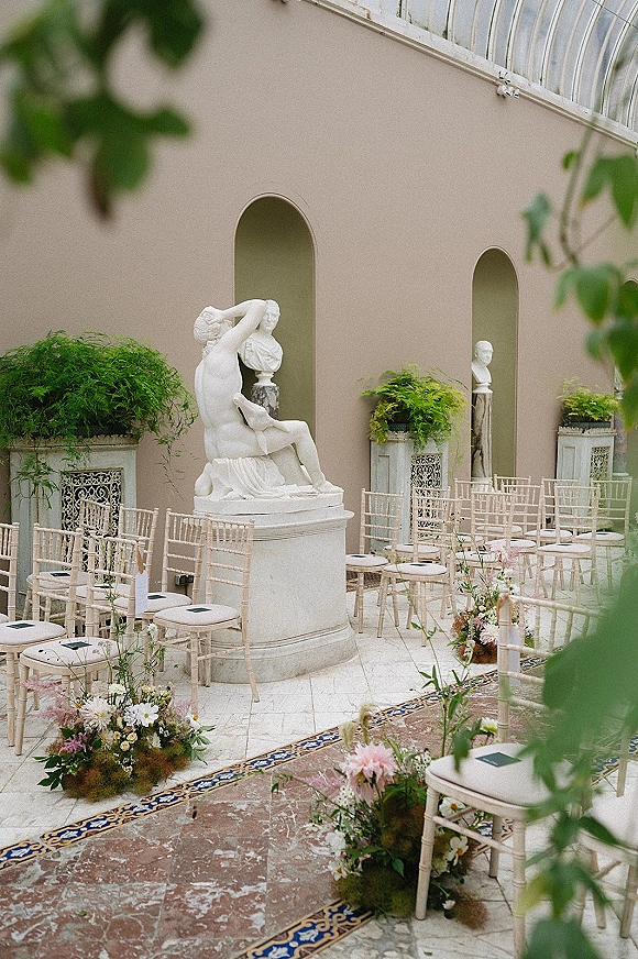 Ceremony setup with outdoor wedding ceremony seating, chiavari chairs lining an aisle runner amid floral clusters in a stone courtyard with arched niches