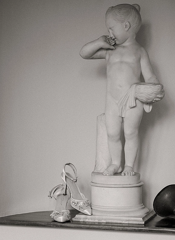 Wedding shoes with embellished bridal heels and ankle straps displayed on a small pedestal statue against a plain wall