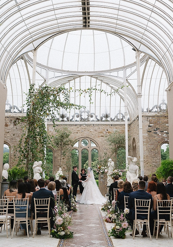 Wedding ceremony with bride and groom at the altar under a greenery arch, pastel aisle florals, in a glass conservatory with arched windows
