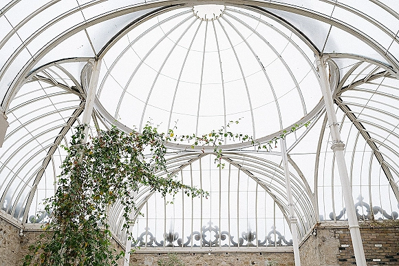 Greenhouse wedding venue with a glass greenhouse ceiling, white metal frame and climbing vines under a domed roof beside brick and stone walls