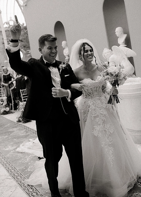 Wedding recessional as newlyweds walk the aisle, groom raising his hand and bride holding bouquet beneath an arched doorway with guests seated
