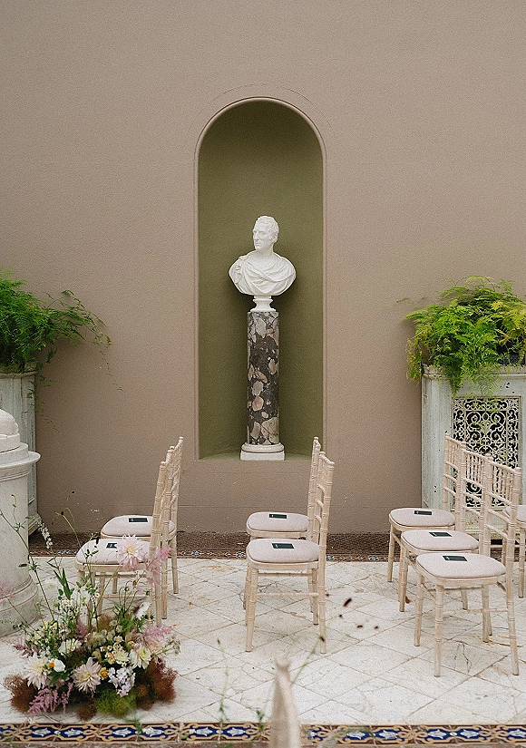 Ceremony setup with outdoor ceremony chairs in neat rows, wood chairs with cushions beside floral arrangement and a classical bust statue in a stucco courtyard