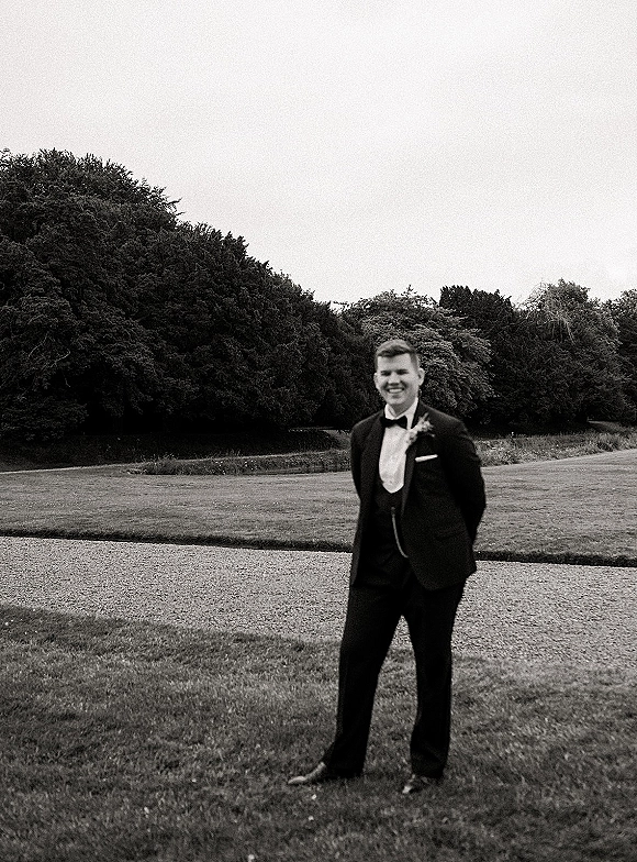 Groom portrait of a smiling groom in black tuxedo with bow tie and boutonniere, hands behind back on a lawn by trees and sky
