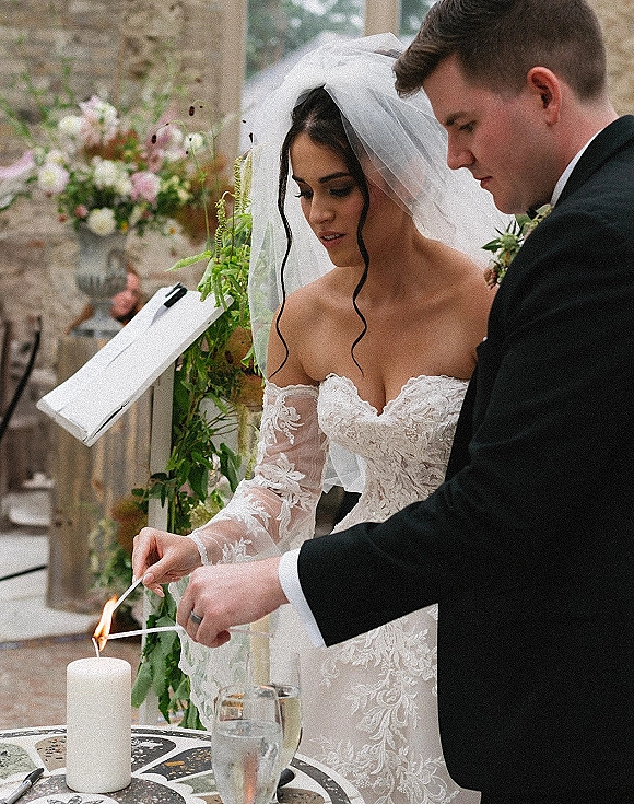 Unity candle ceremony as bride and groom light taper candles, her lace off-shoulder sleeves and veil by a stone patio wall