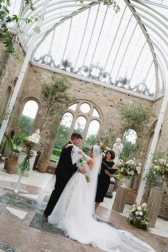 Wedding ceremony moment as bride and groom stand at the altar with veil and lace sleeves, under a glass conservatory ceiling with florals