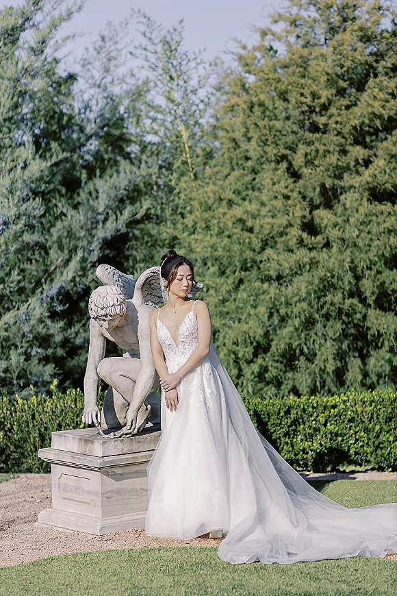 Bridal portrait of a bride in a sparkly spaghetti strap gown with a long tulle train beside a stone angel statue in a garden
