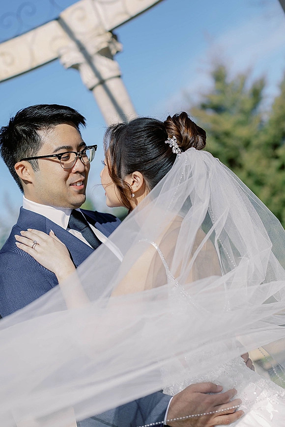 Couple portrait of bride and groom embrace, her veil blowing as they lean forehead to forehead by a stone cross under blue sky
