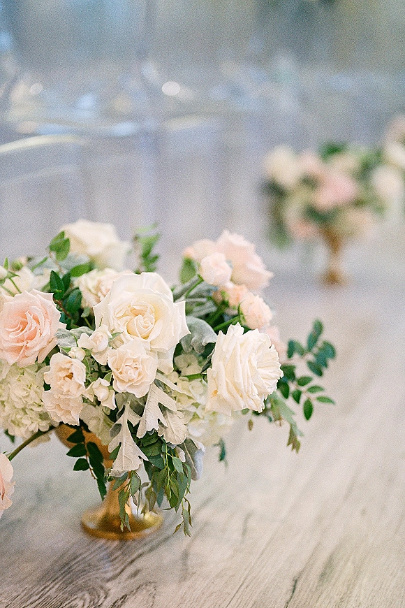 Wedding centerpiece in a gold compote centerpiece vase with blush and white roses and eucalyptus on a reception table with chairs behind