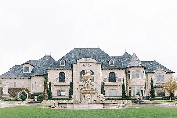 Wedding venue exterior of a castle-like estate wedding venue with a stone fountain, ivy-covered facade, and manicured hedges under an overcast sky