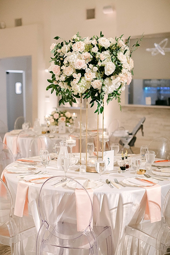 Reception tablescape with tall floral centerpiece on a gold stand, blush napkins, and votive candles in a bright indoor reception room
