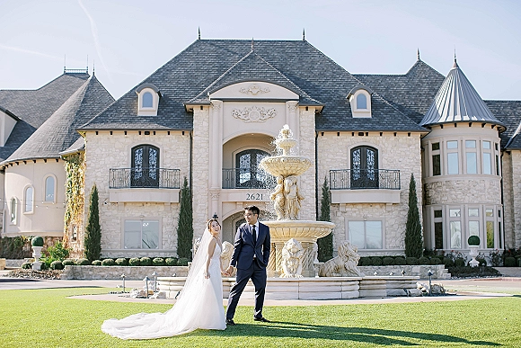 Couple portrait of bride and groom holding hands on a lawn, her long veil and dress train flowing near a fountain by a stone mansion