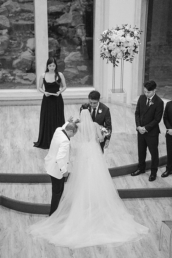 Ceremony moment as bride and groom stand at the altar during wedding ceremony prayer, bride holding bouquet before large windows indoors