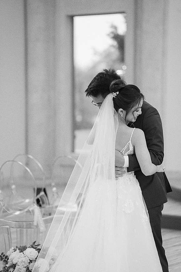 First dance as bride and groom dancing, her long veil trailing over a sparkly gown while he holds her in an indoor reception by a window