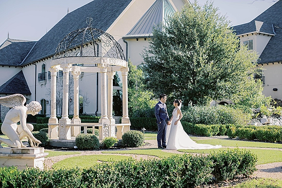 Couple portrait of bride and groom holding hands, facing each other, her long train and veil flowing on a garden lawn by a gazebo pavilion