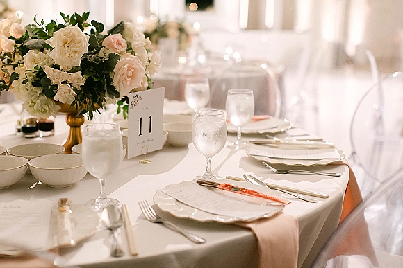 Reception tablescape with wedding table setting, rose and greenery centerpiece in a gold compote vase, menus, place cards, and votive candles in a softly lit room
