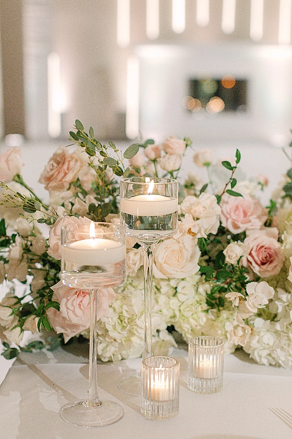Reception centerpiece with wedding flower centerpiece of blush roses and white blooms, greenery, and floating candles in glass vases on a tabletop