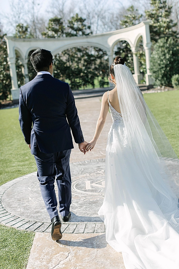 Wedding couple walking hand in hand, bride and groom walking away with cathedral veil and lace gown on a stone path by white arches
