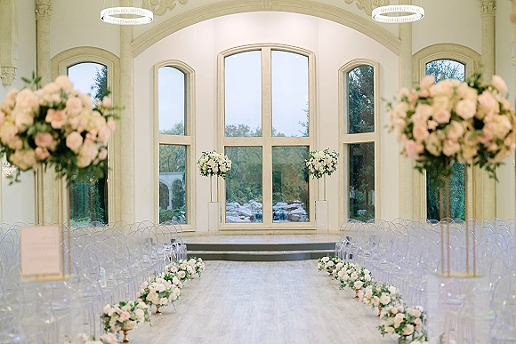 Indoor ceremony setup with a wedding ceremony aisle lined in white and blush florals, clear chairs, and arched windows behind the altar