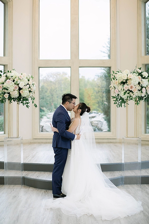 Wedding kiss portrait of bride and groom kissing beneath a bridal veil on stone steps, framed by floral stands and large windows with trees outside