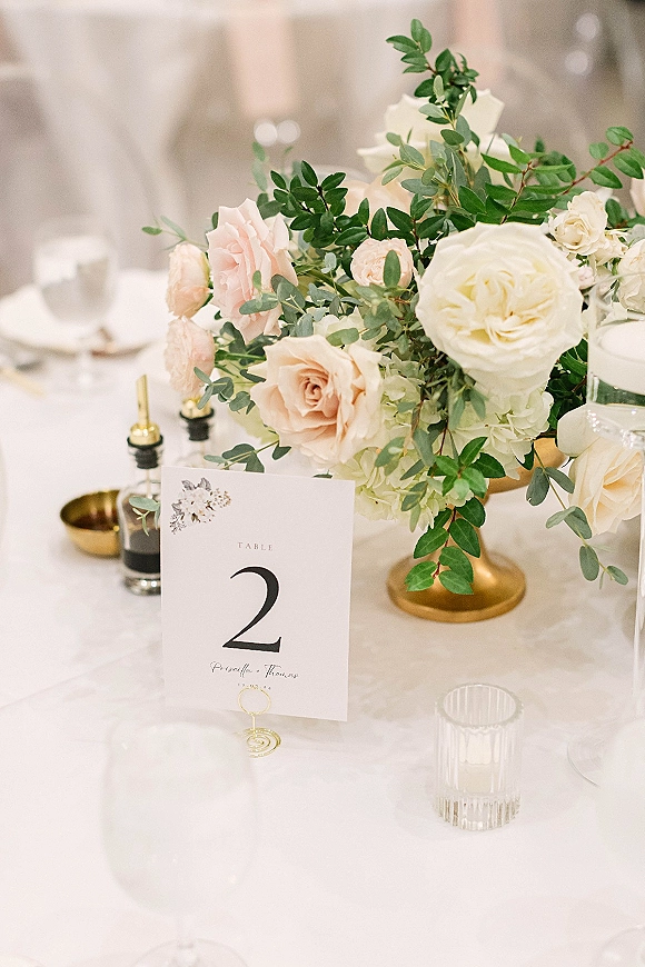 Reception tablescape with wedding table centerpiece of roses and greenery in a gold compote vase, table number card on white linen, chairs blurred behind
