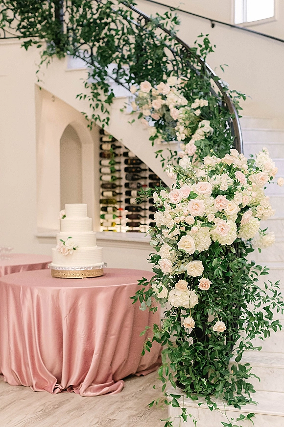 Wedding cake display with a three-tier white cake on a stand, set on blush satin tablecloth beneath a rose and hydrangea arch indoors