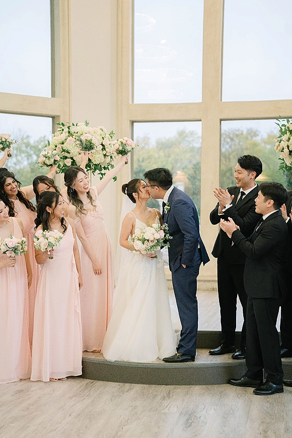 Wedding kiss as bride and groom kiss on a raised indoor stage, wedding party cheers with blush bouquets by large windows and trees outside