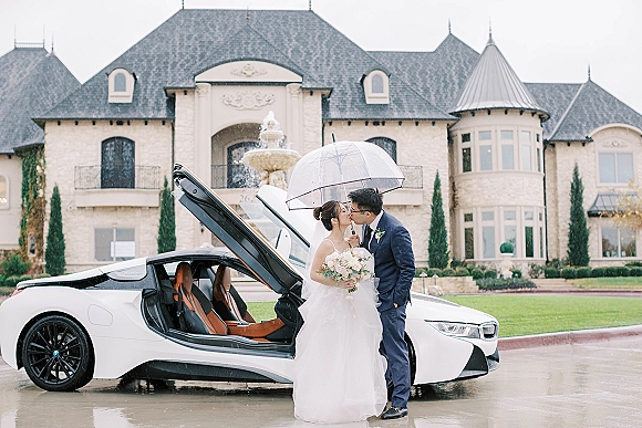 Wedding kiss portrait of bride and groom kissing under umbrella, beside a white convertible on a rainy mansion driveway near a fountain