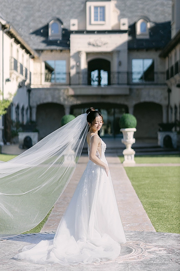 Bridal portrait of a bride in a wedding dress with a long veil blowing in the wind on a stone walkway by a manor exterior