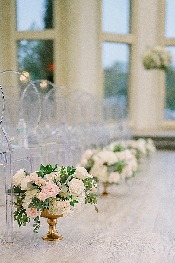 Ceremony aisle decor with aisle floral arrangements of blush and white roses on gold urns beside clear acrylic chairs in a window-lit venue