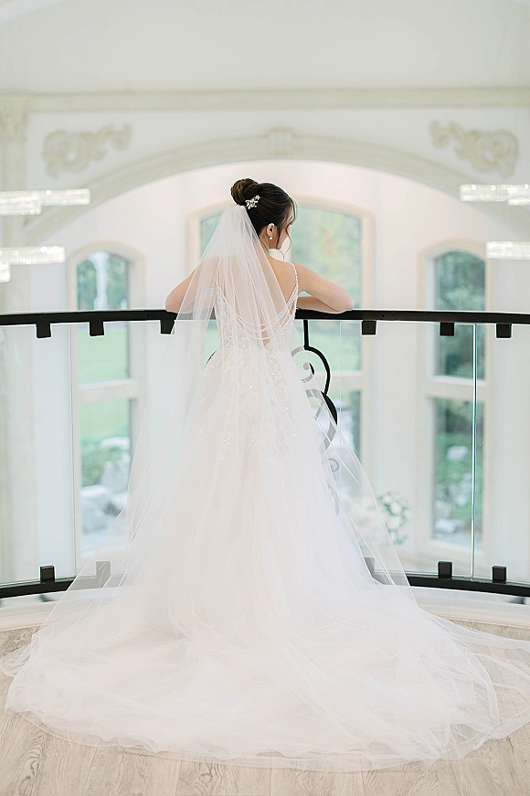Bridal portrait from behind showing a long cathedral veil and low-back wedding dress, bride leaning on glass railing by arched windows