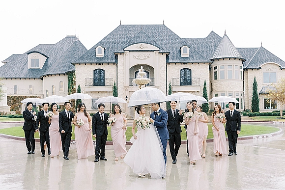 Wedding party portrait with bride and groom under umbrella, holding bouquets on wet pavement by a stone mansion and fountain in rain