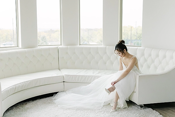 Bridal portrait of a bride putting on heels, seated on a tufted white sofa in a tulle gown with jeweled hairpiece by bright windows