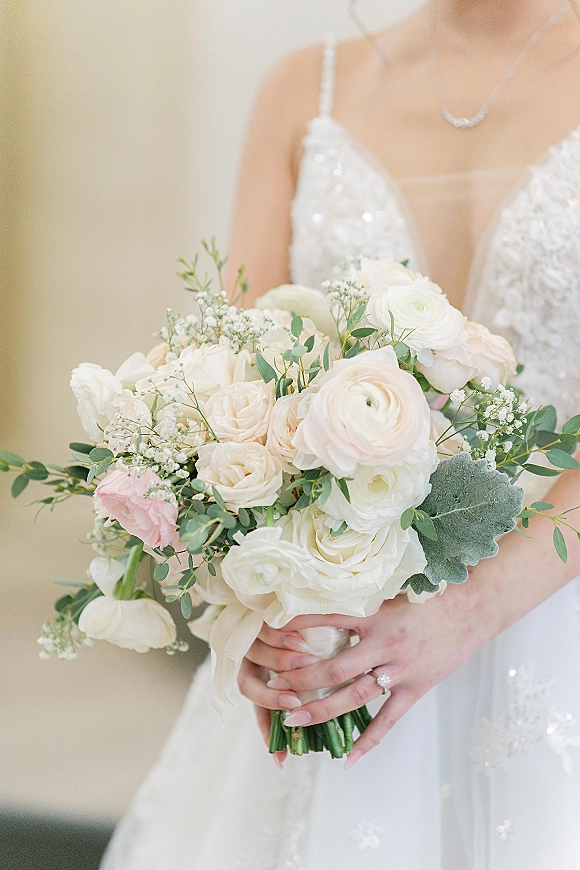 Bridal bouquet of white roses and ranunculus with eucalyptus, held against a sequin wedding dress, engagement ring visible on hand