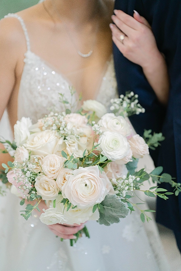 Bridal bouquet of blush and white flowers with greenery and baby's breath held against a lace bridal gown, with neutral wall backdrop